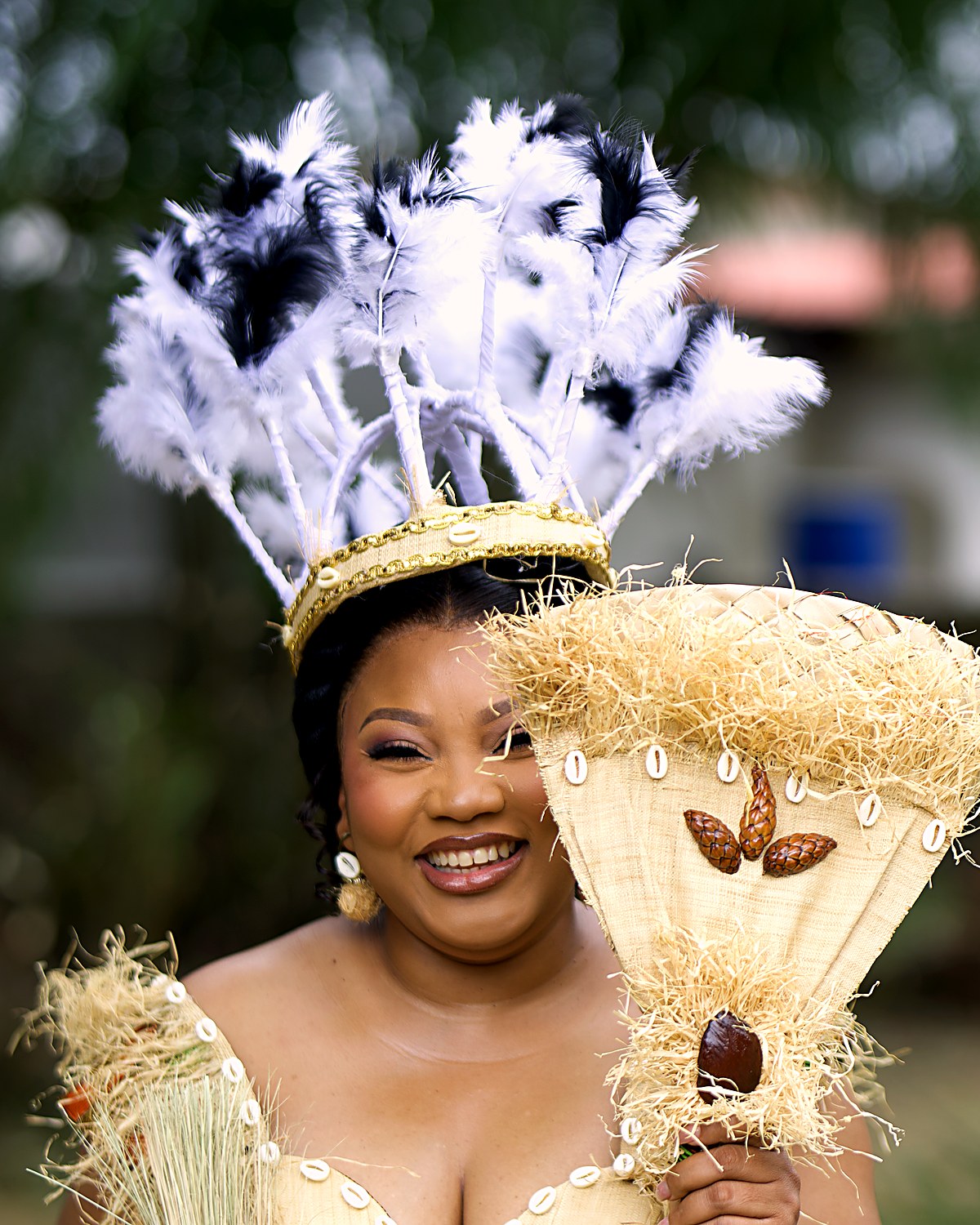 Rhudye en tenue de mariage coutumier : robe de raphia et cauris, couronne de plumes, éventail